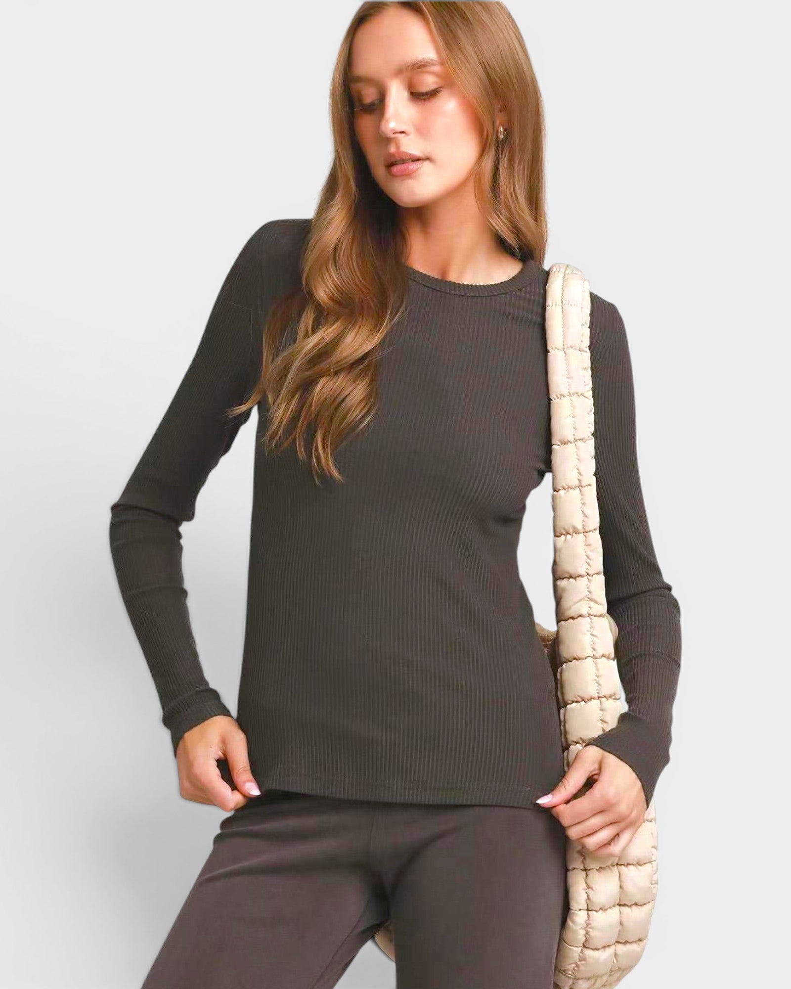 Woman wearing a dark gray long-sleeve top and pants, holding a beige bag against a light gray background.