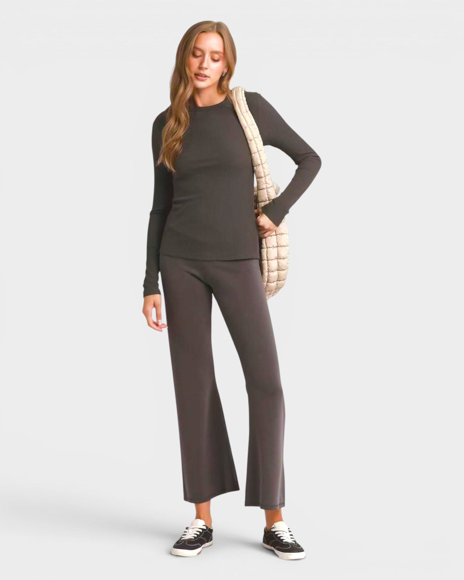 Woman wearing a dark gray long-sleeve top and wide-leg pants, holding a beige bag on a light gray background.
