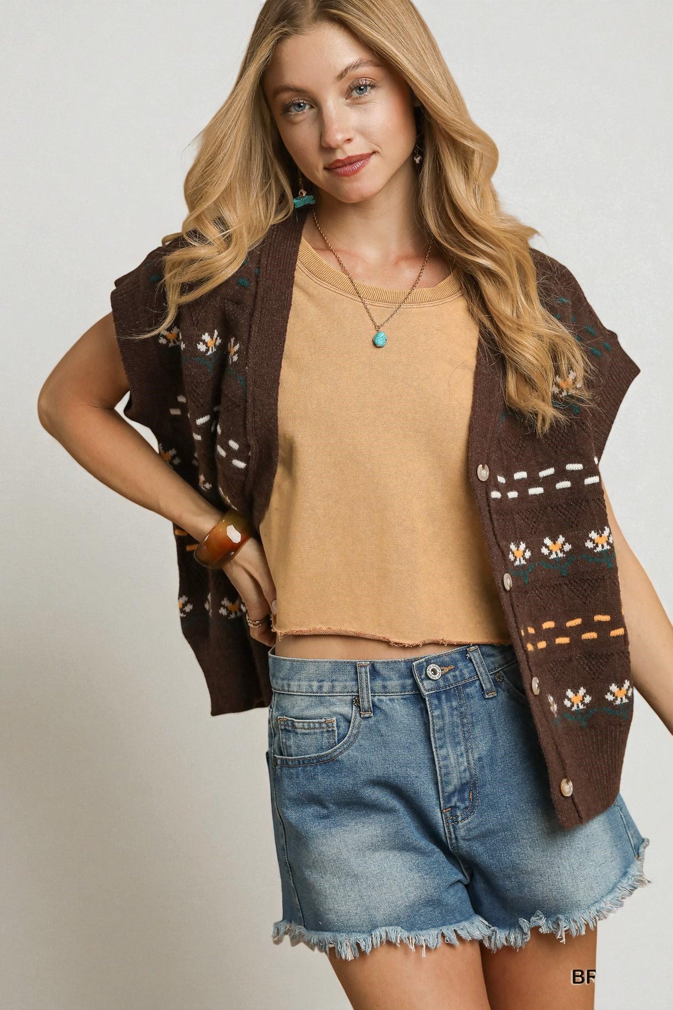 Woman wearing a brown patterned vest over a beige top and denim shorts on a plain background