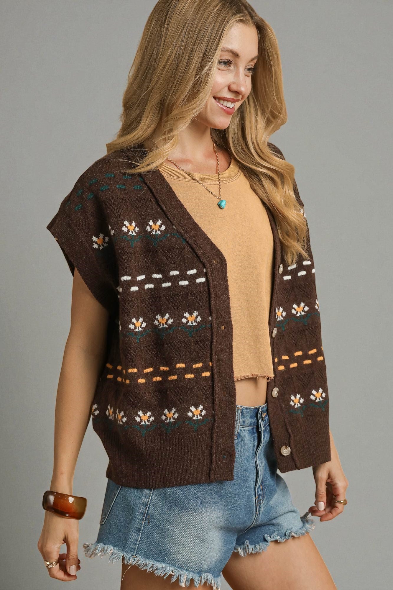 Woman wearing a brown patterned cardigan over a beige top and denim shorts on a gray background