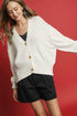 Woman wearing a white cardigan with gold buttons and black shorts on a red background