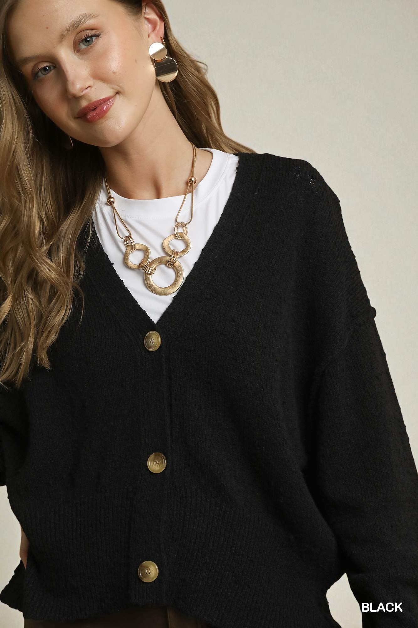 Woman wearing a black cardigan over a white shirt with gold buttons, gold necklace, and earrings.