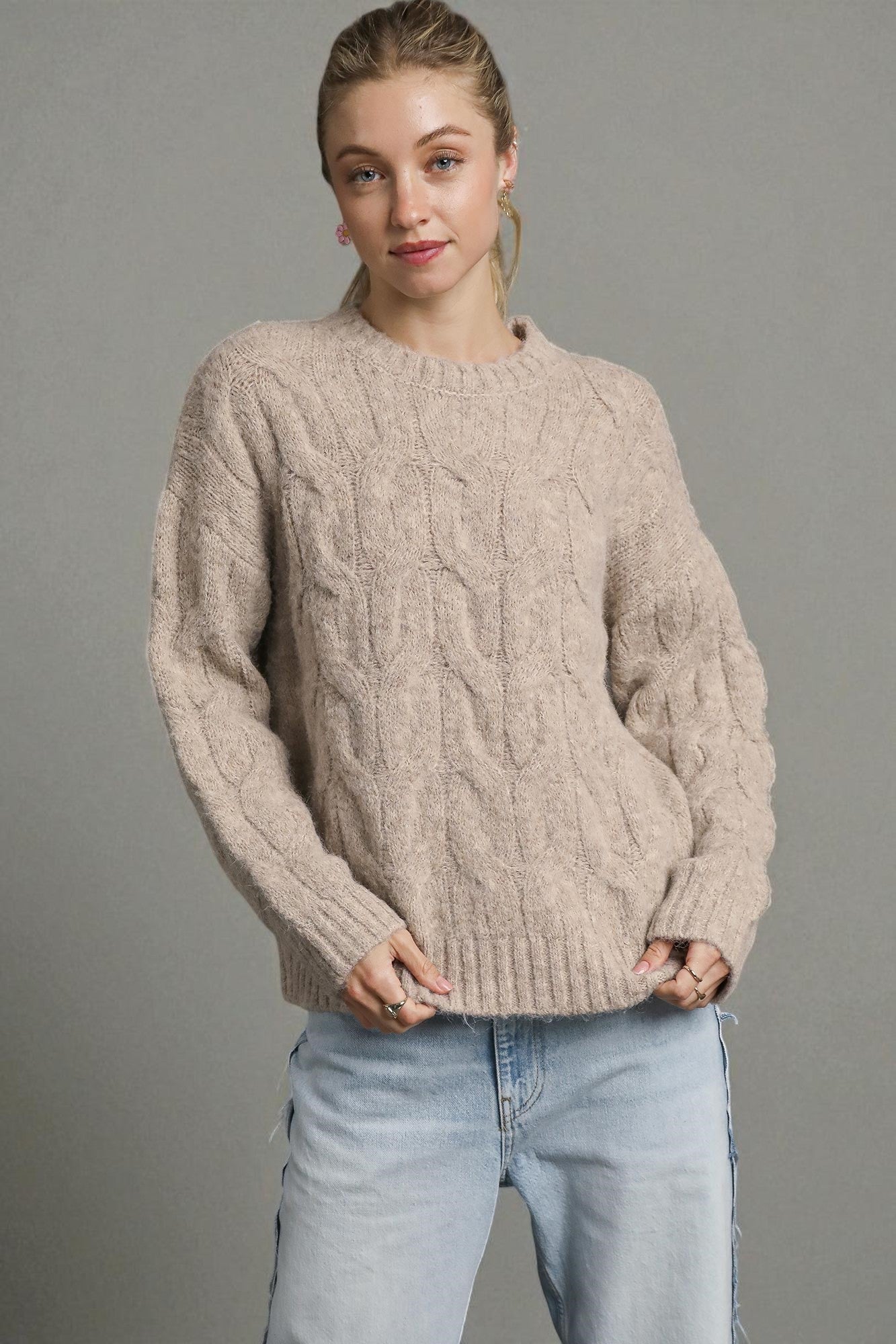 Woman wearing a beige cable knit sweater and light blue jeans against a gray background
