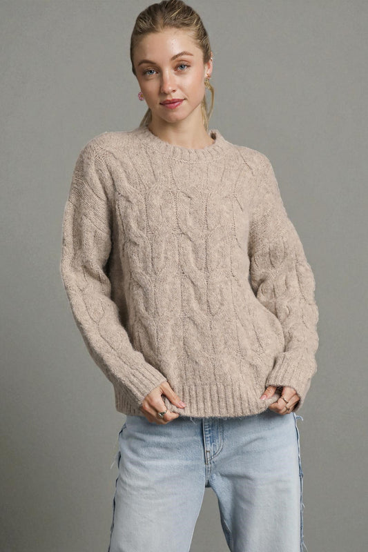 Woman wearing a beige cable knit sweater and light blue jeans against a gray background