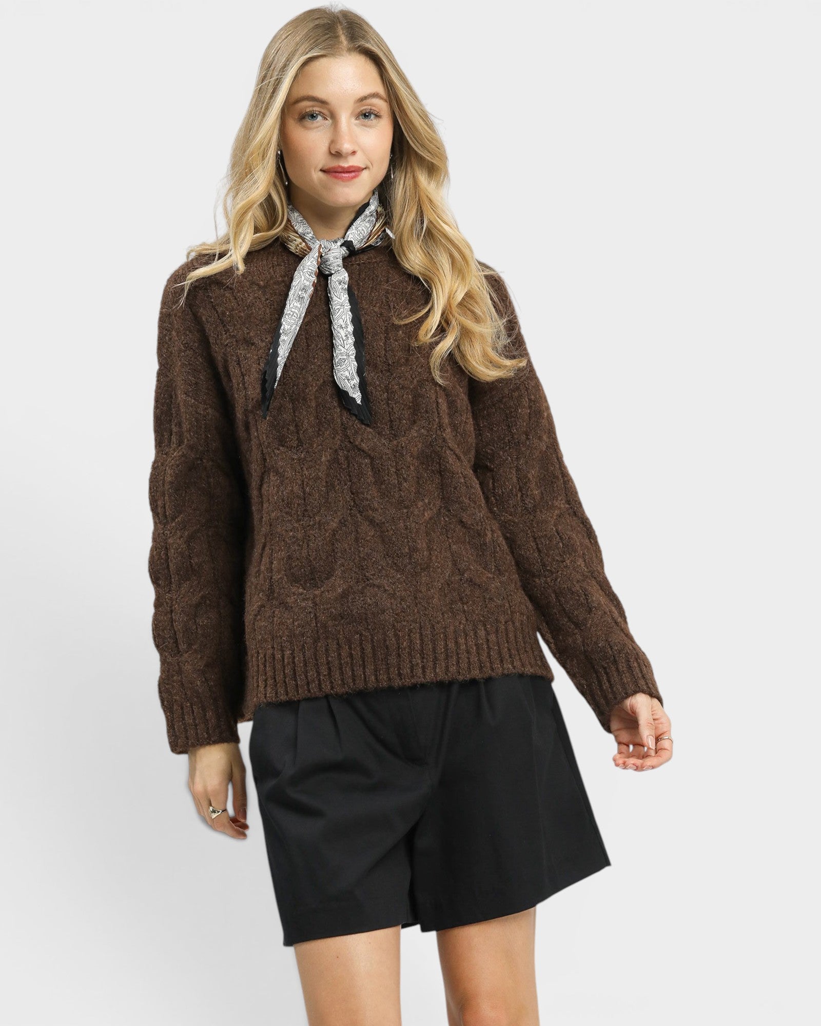 Woman wearing a brown cable knit sweater with a black and white scarf, standing against a plain background.