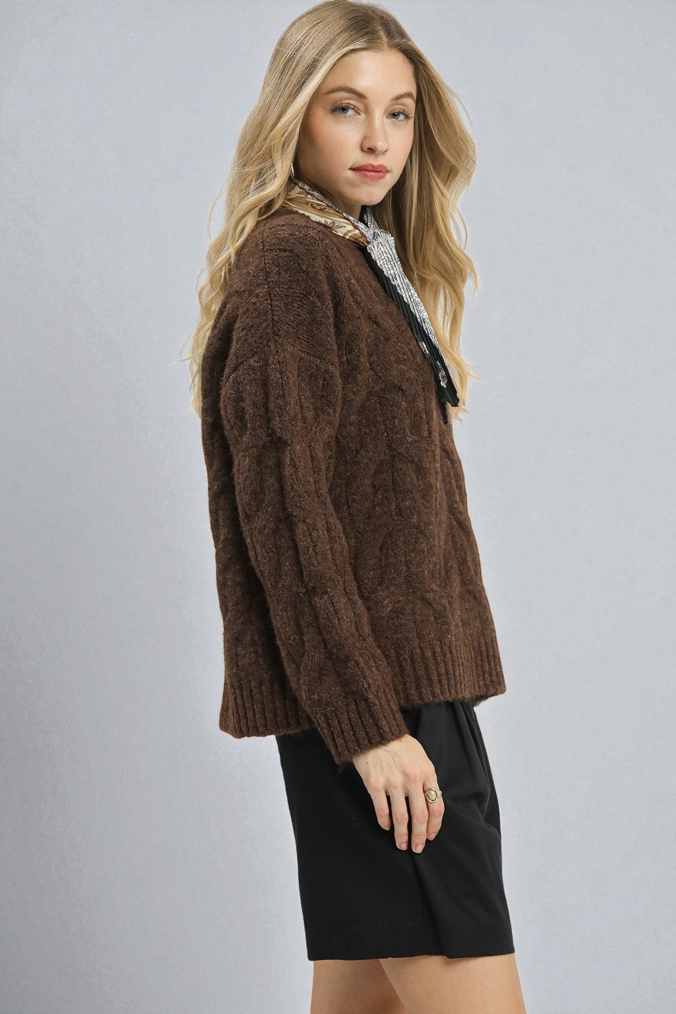 Woman wearing a brown cable knit sweater over a patterned shirt on a gray background