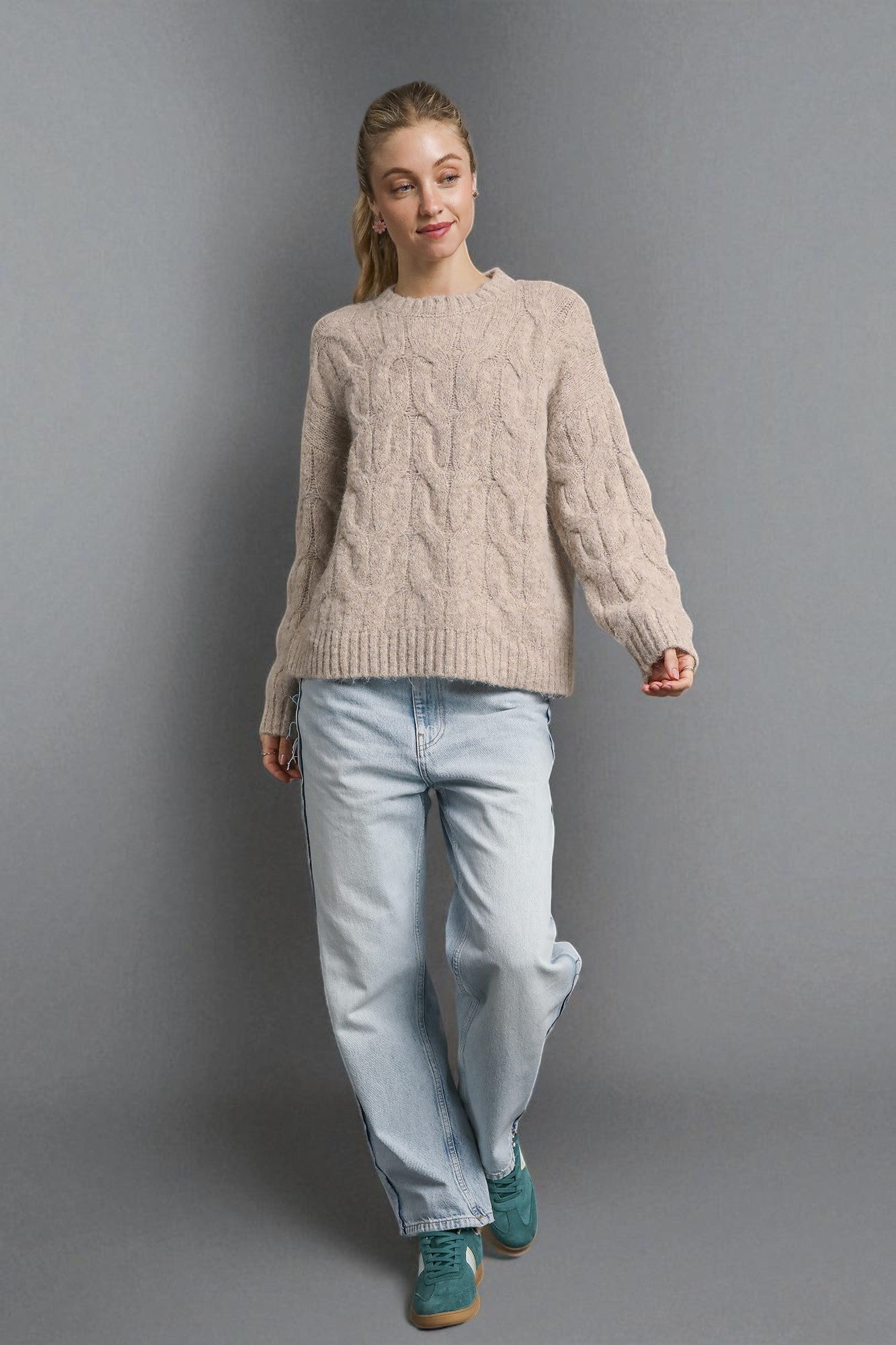 Woman wearing a beige cable knit sweater and light blue jeans against a gray background