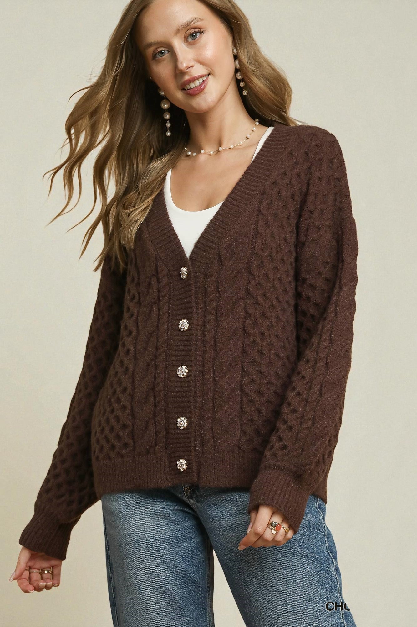 Woman wearing a brown cable knit cardigan over a white top and blue jeans on a beige background