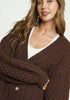 Woman wearing a brown cable knit cardigan over a white top on a light background
