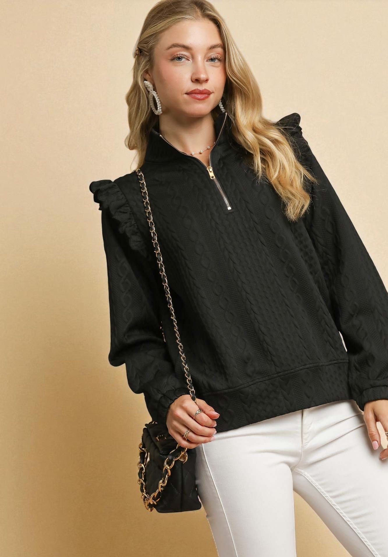 Woman wearing a black pullover with a zipper, white pants, and holding a black handbag with a gold chain on a beige background.