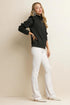 Woman wearing a black top with ruffled sleeves and white pants against a beige background