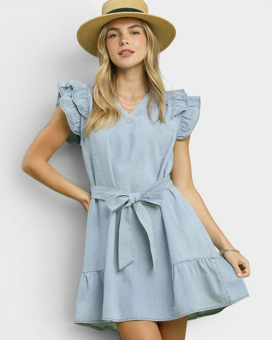 Woman wearing a light blue dress with ruffled sleeves and a belt, standing against a plain background.