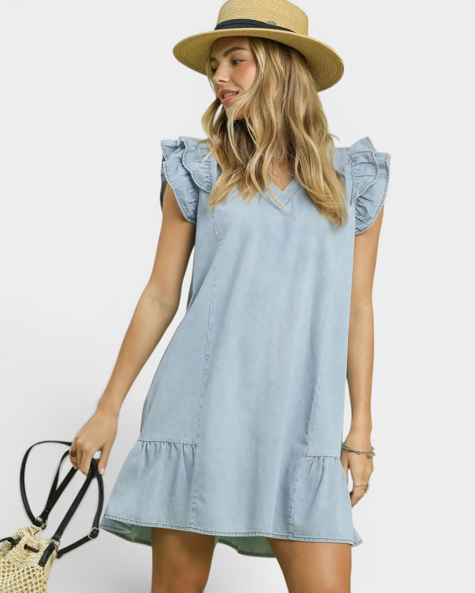 Woman wearing a light blue denim dress with ruffled sleeves and bottom hem, holding a straw bag against a white background.