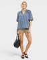 Woman wearing a blue blouse and beige shorts, holding a black handbag on a white background