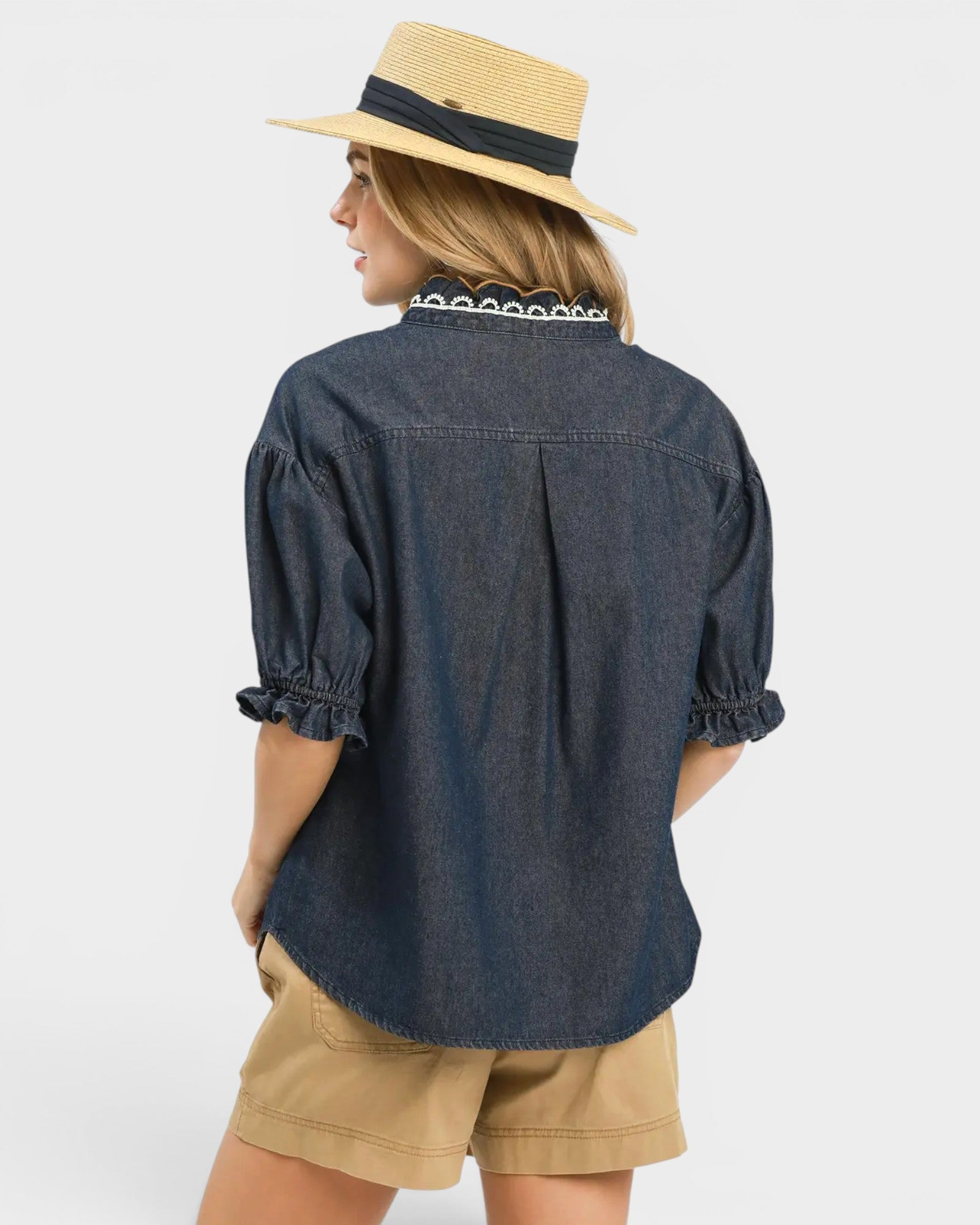 Person wearing a navy denim shirt and beige shorts with a straw hat on a white background