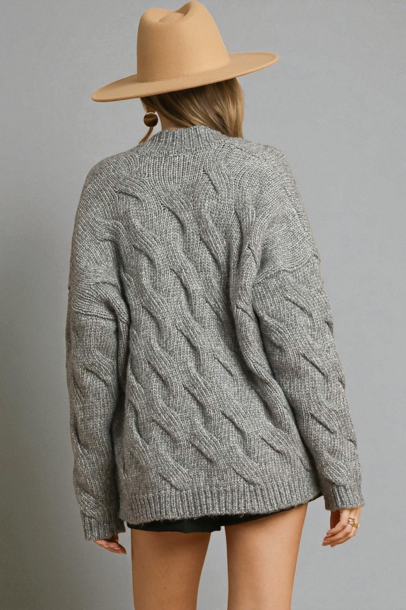 Person wearing a gray cable knit sweater and tan hat on a gray background