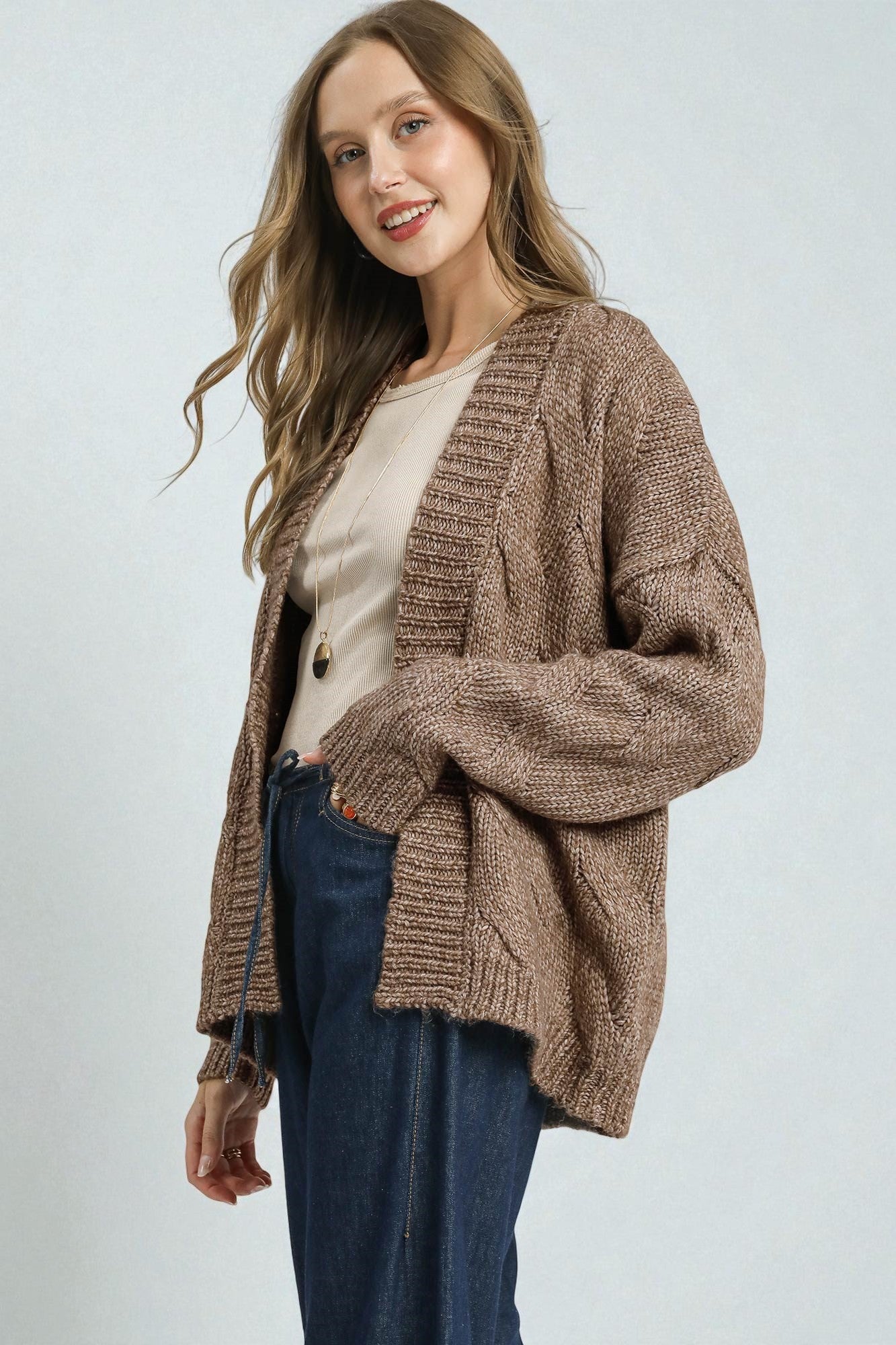 Woman wearing a brown knitted cardigan over a white shirt and blue jeans on a light gray background