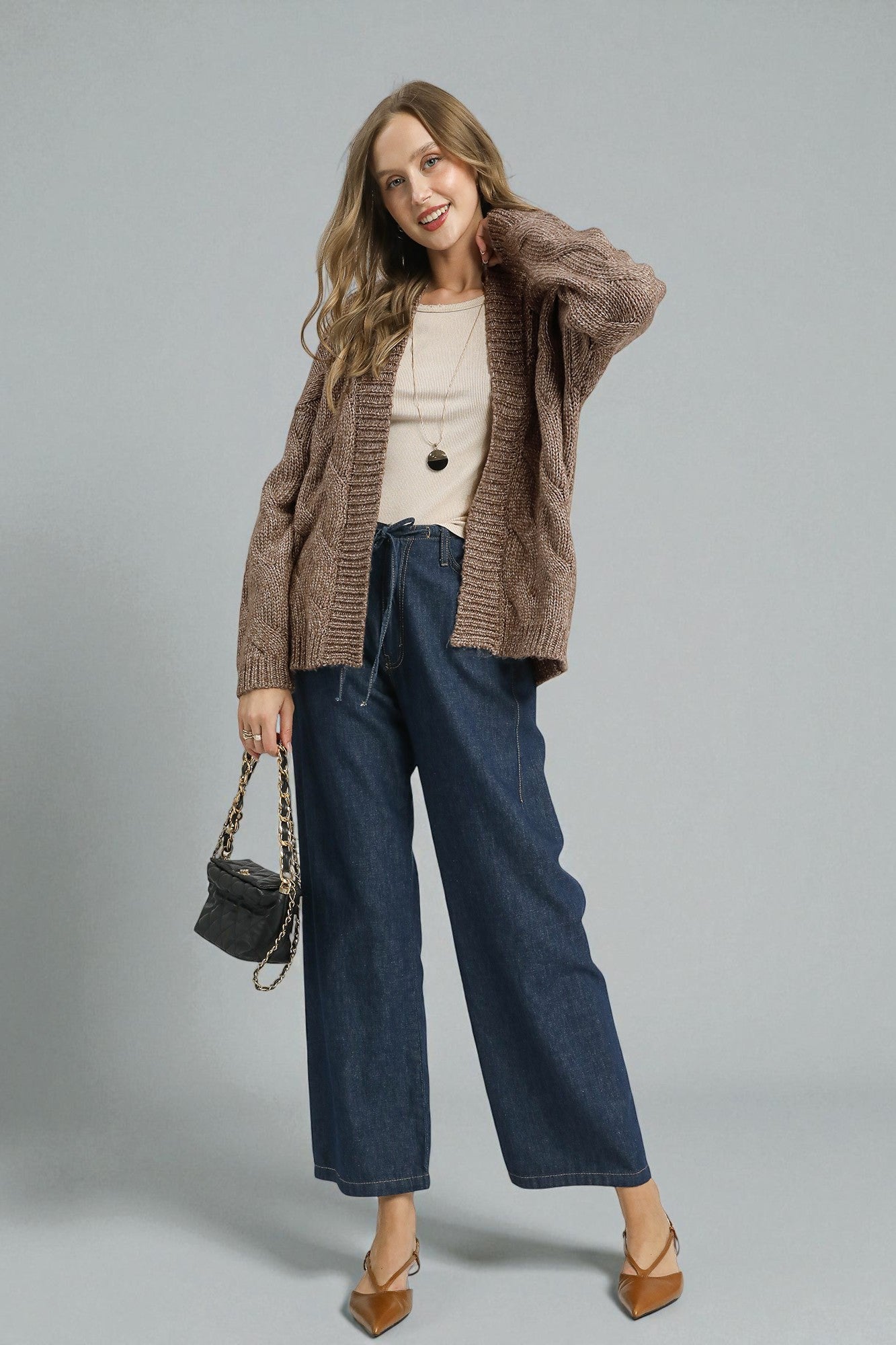 Woman wearing a brown cardigan, beige top, blue jeans, and carrying a black handbag on a gray background