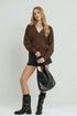 Woman wearing a brown cardigan, black skirt, and black boots holding a black handbag on a white background