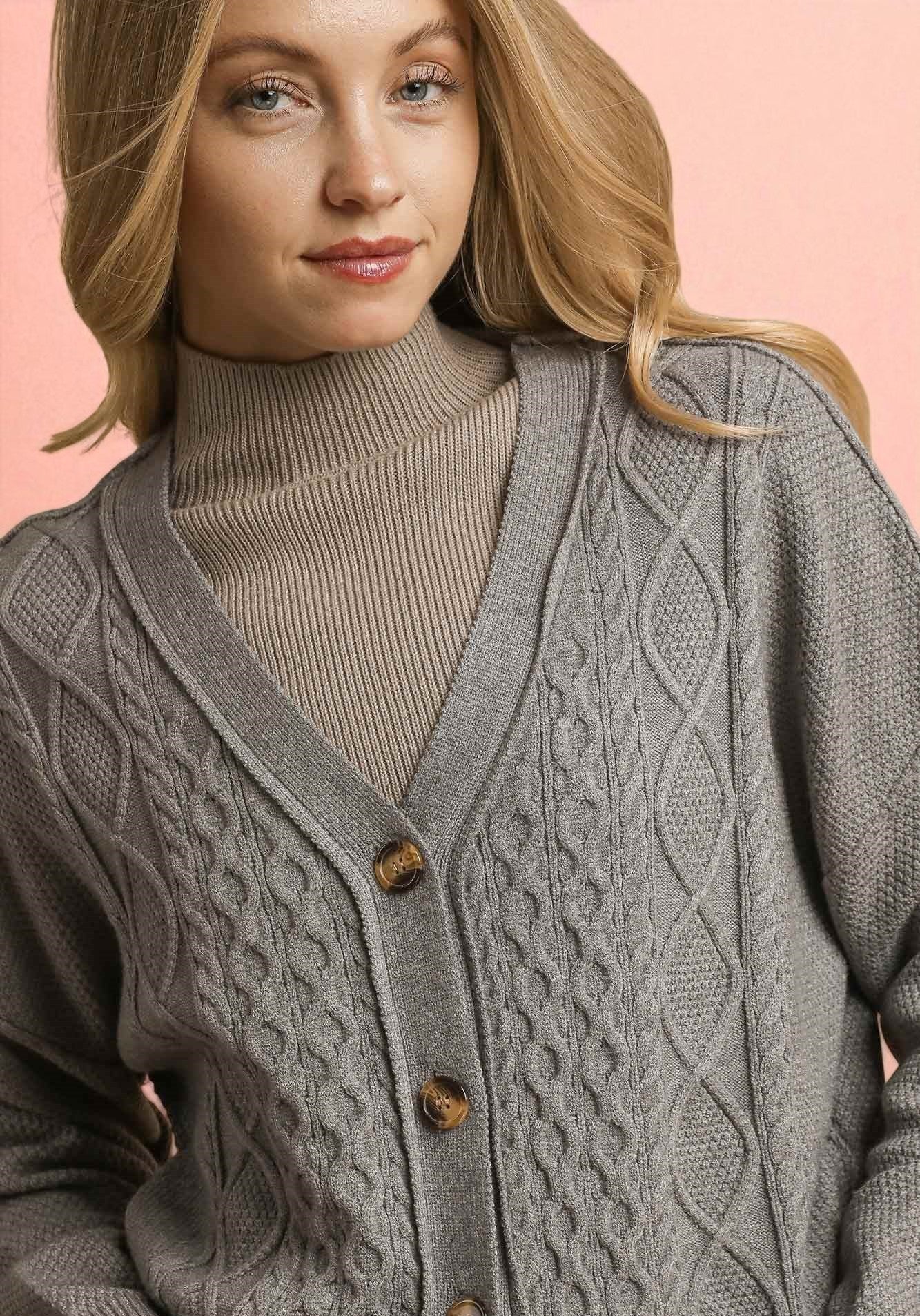Woman wearing a gray cable knit cardigan over a beige turtleneck against a pink background