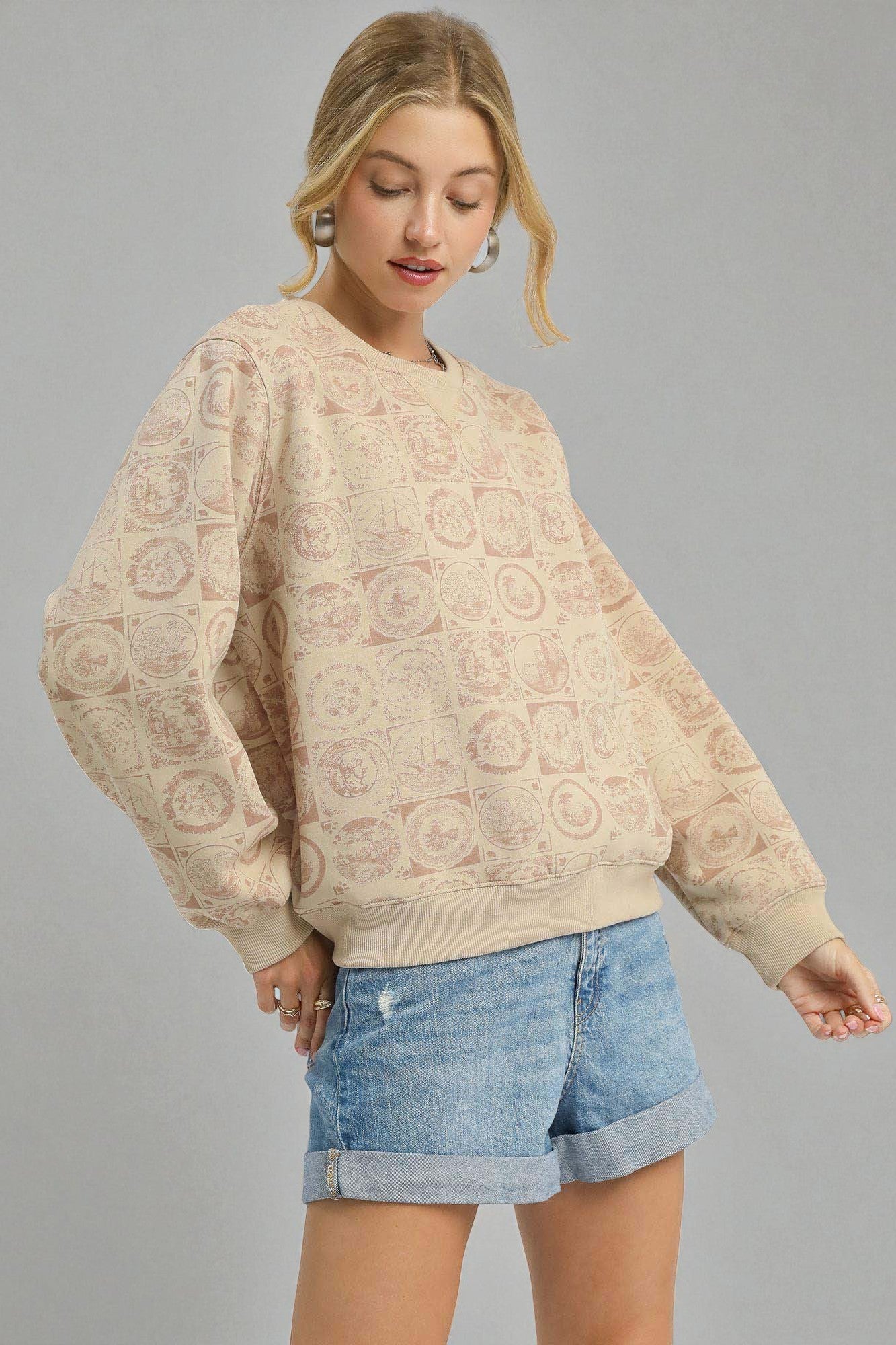 Woman wearing a beige patterned sweater and denim shorts on a gray background