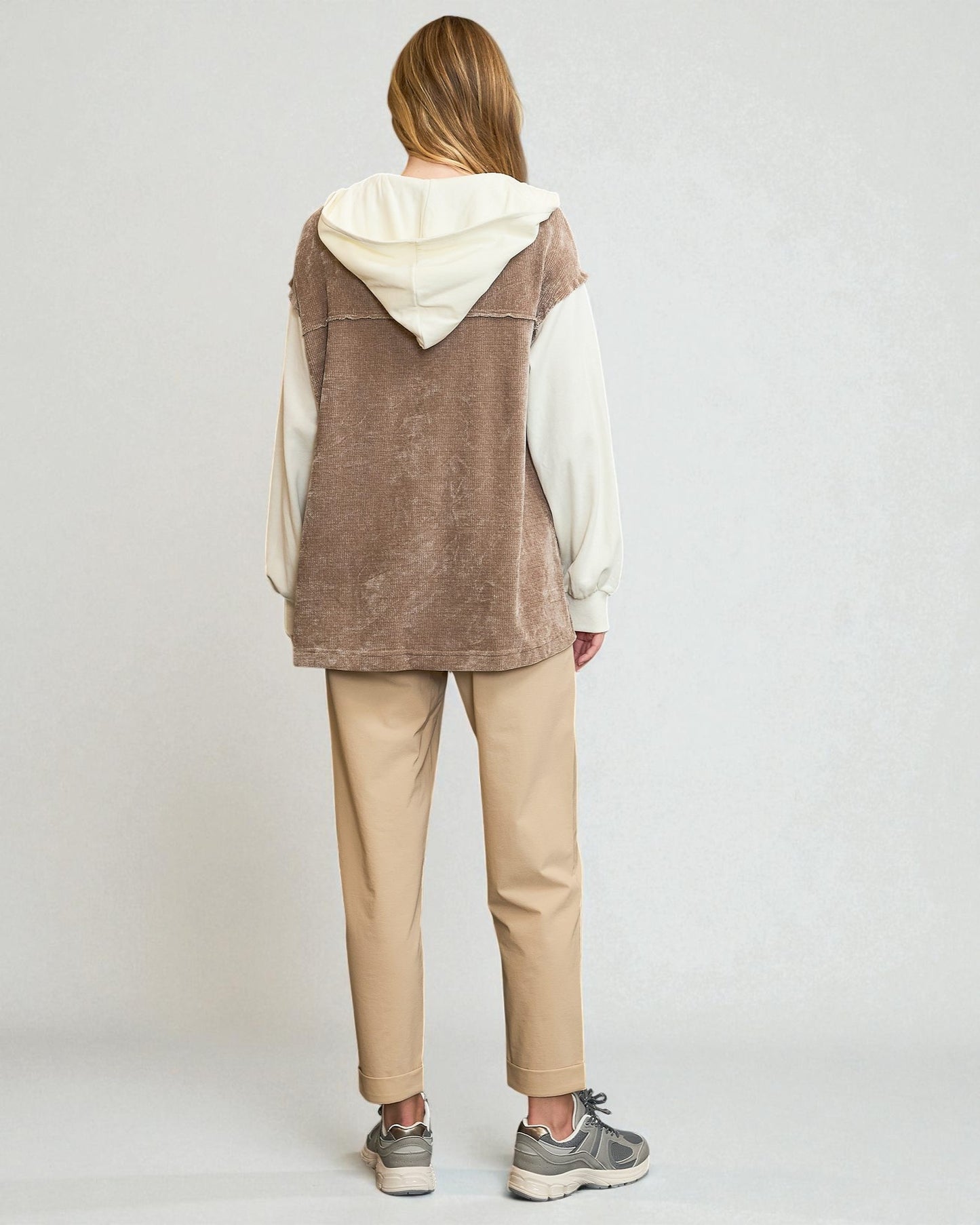 Person wearing a brown vest over a white hoodie with beige pants on a white background