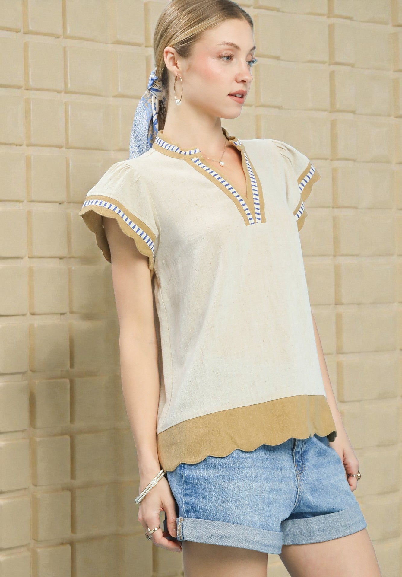 Woman wearing a beige top with scalloped hem and denim shorts against a tiled wall.