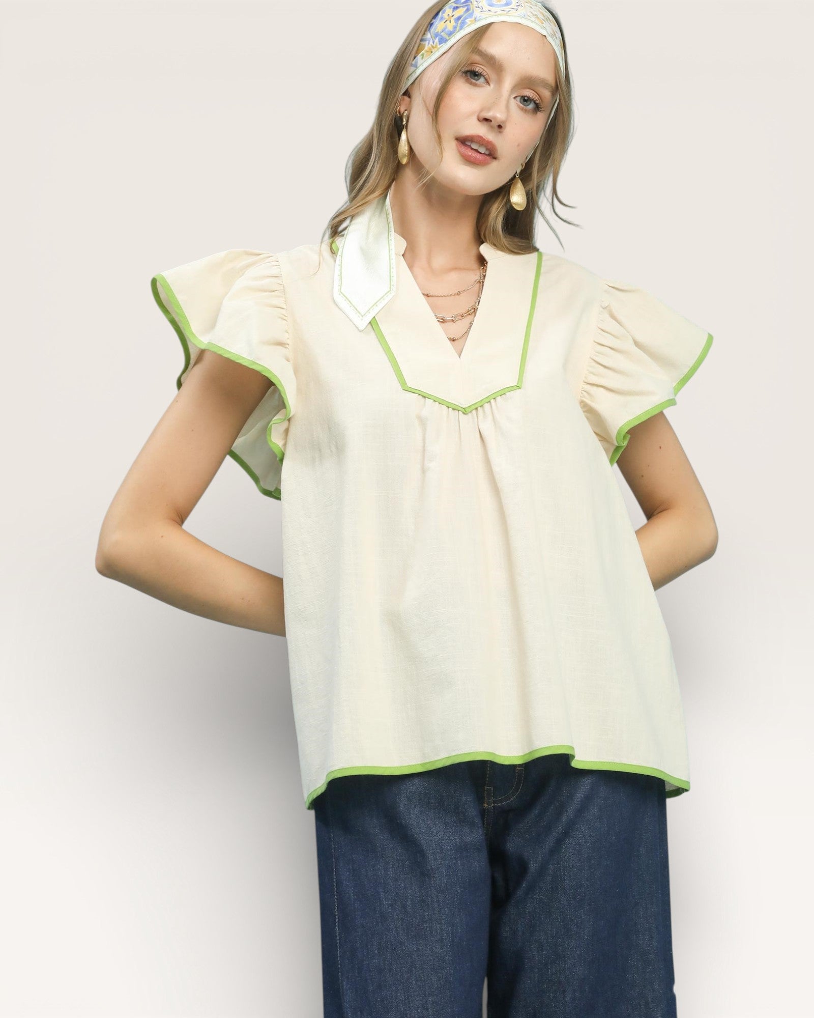 Woman wearing a light-colored blouse with green trim and blue pants on a plain background