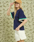 Woman wearing a navy blue shirt with green trim and white shorts, standing against an abstract beige wall.