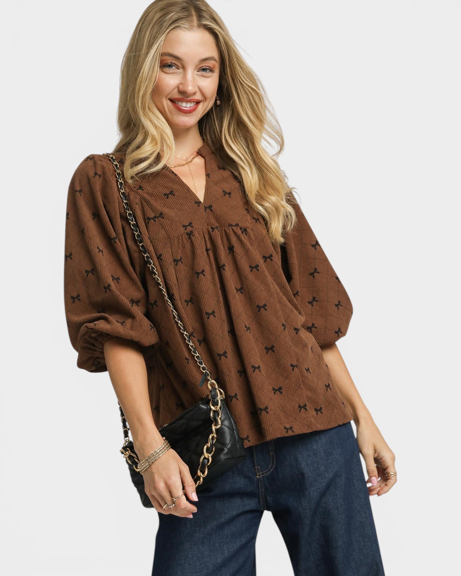 Woman wearing a brown blouse with black patterns and blue jeans, holding a black handbag.
