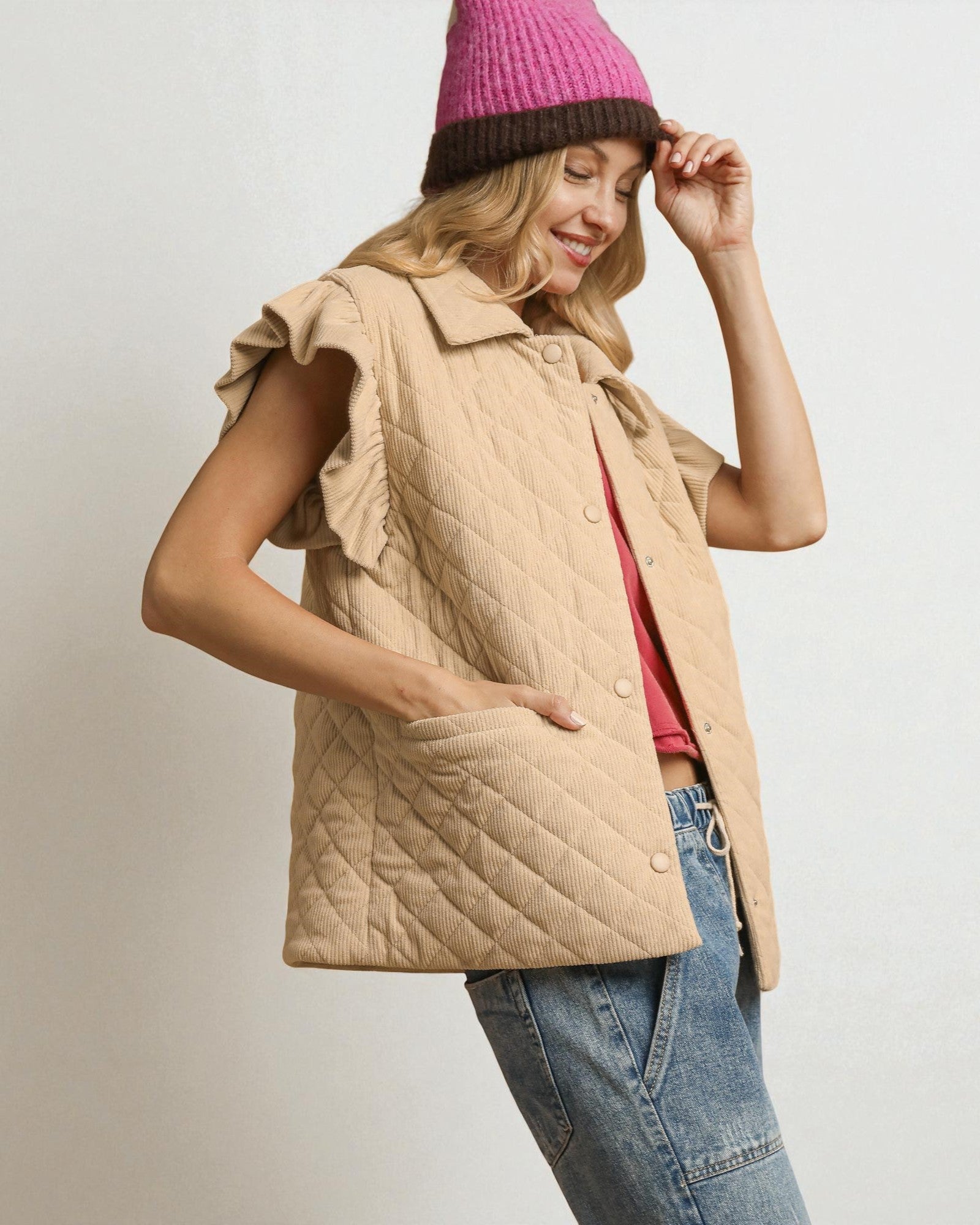 Woman wearing a beige quilted vest, pink beanie, and blue jeans on a plain background