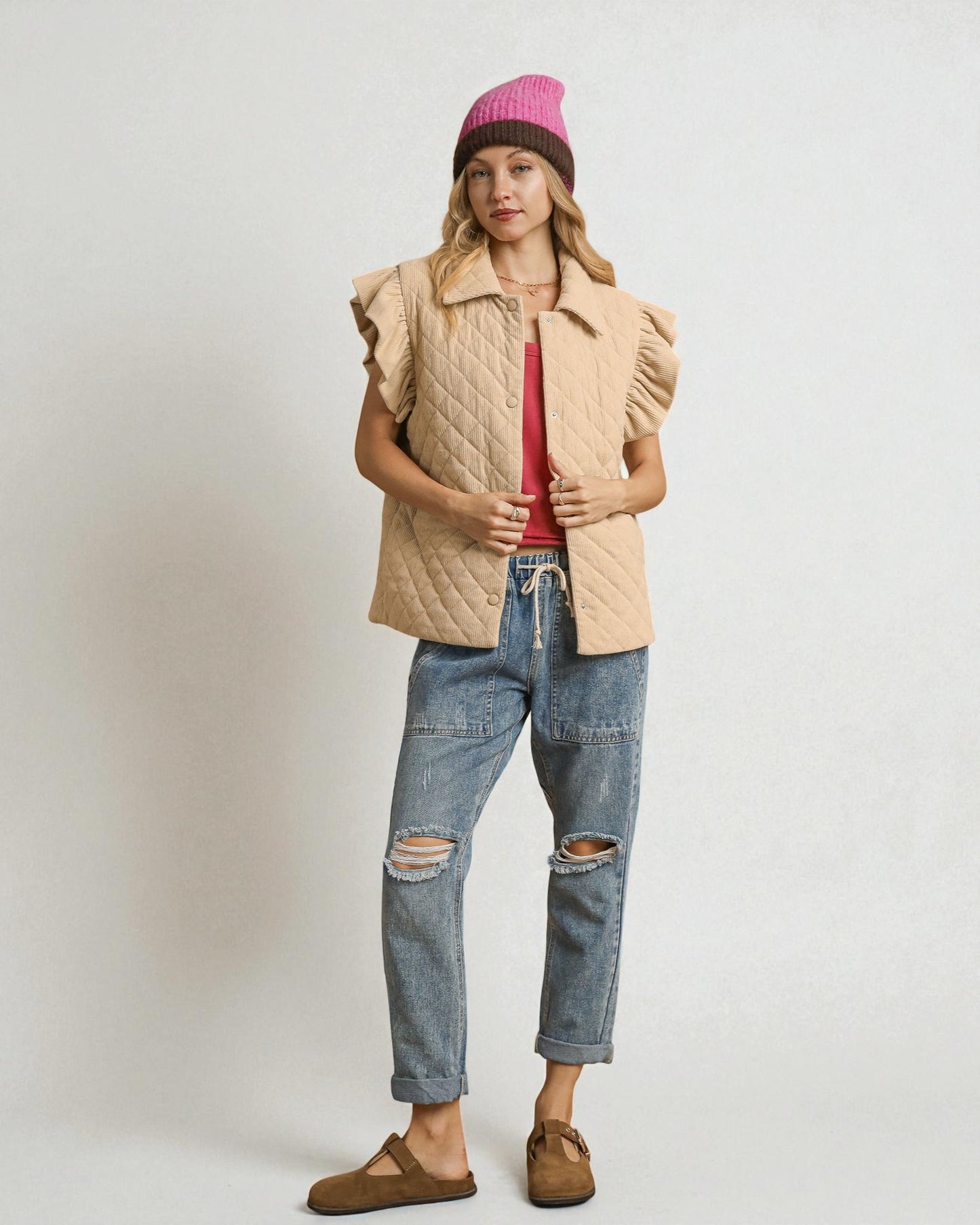 Person wearing a beige quilted vest, pink beanie, red shirt, blue jeans, and brown shoes on a white background