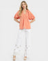 Woman wearing a coral blouse and white pants with floral embroidery on a white background