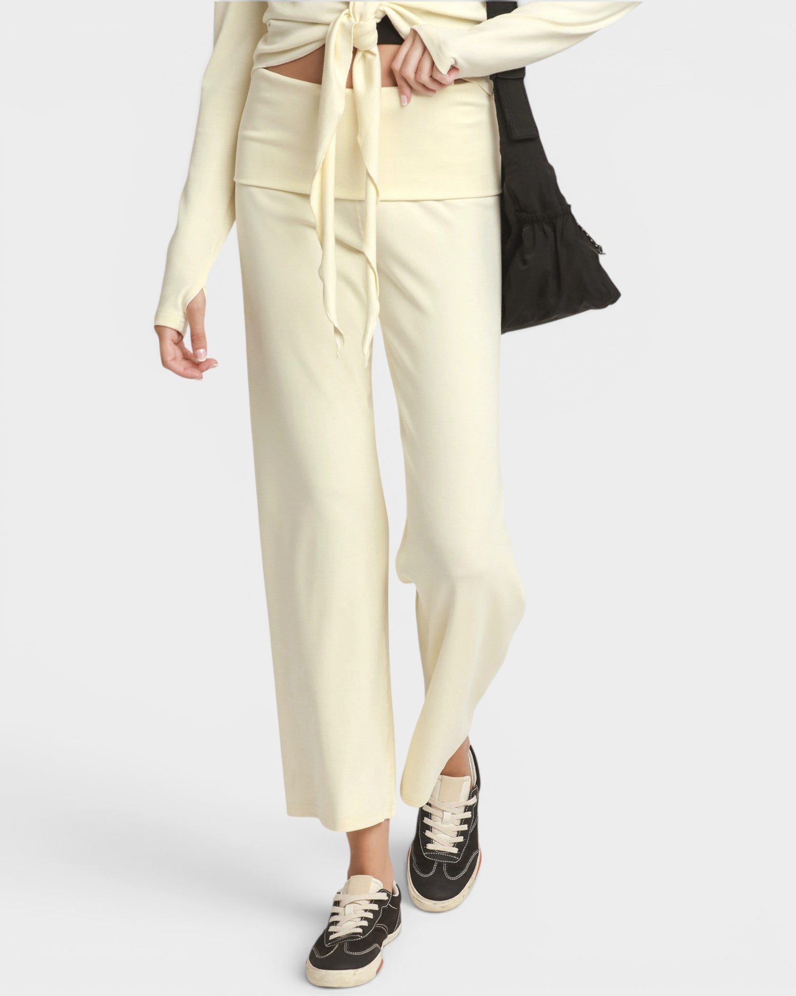 A model wearing cream wide-leg cropped pants with a matching tie-front waistband, paired with black sneakers and a black shoulder bag, against a light gray studio background.