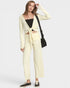 A woman modeling a cream-colored two-piece lounge set consisting of a long-sleeve wrap cardigan and matching wide-leg pants, styled with a black crop top, black bag, and black sneakers against a light gray background.