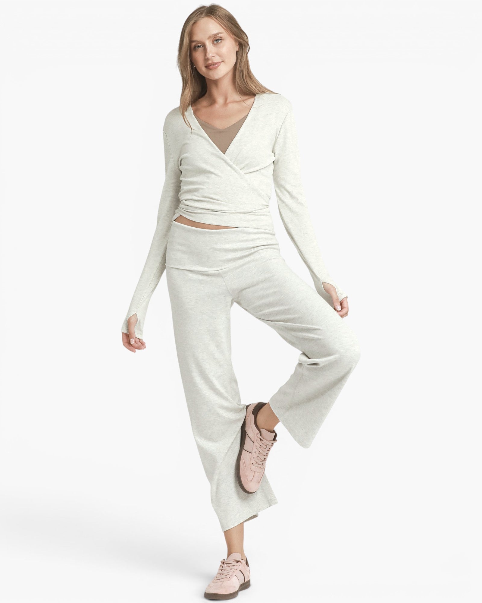 A woman modeling a light cream knit loungewear set consisting of a long-sleeve wrap-style top and matching wide-leg pants, paired with light pink sneakers against a plain white studio background.