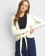 A woman modeling a cream-colored open-front cardigan tied at the waist over a black top and navy blue drawstring pants, set against a plain light gray studio background.