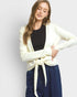 A woman modeling a cream-colored open-front cardigan tied at the waist over a black top and navy blue drawstring pants, set against a plain light gray studio background.
