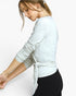 A woman wearing a light heather gray long-sleeve top with ruched sleeves and a side tie detail at the waist, paired with black leggings, shown against a plain light gray background.