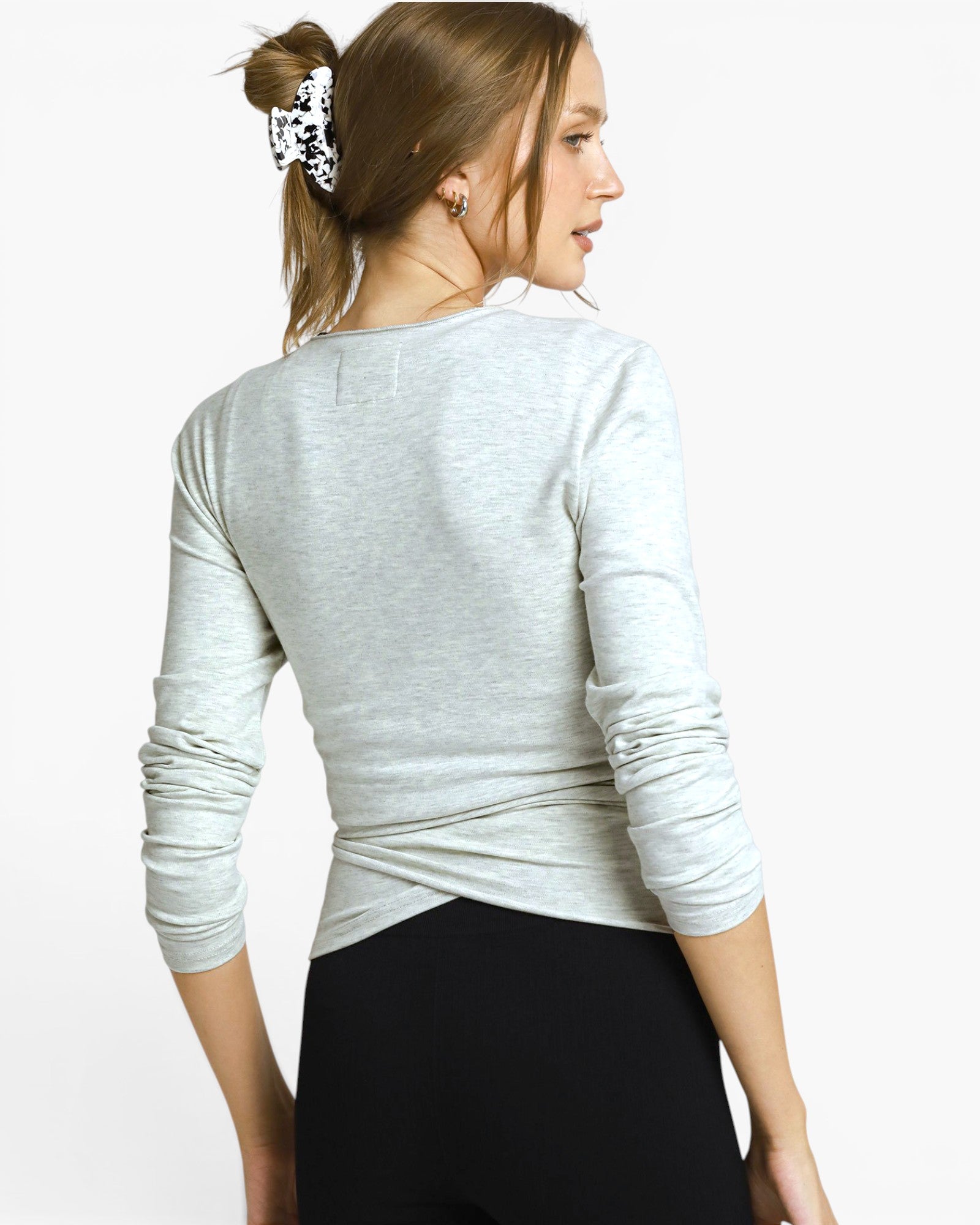 A woman shown from the back wearing a light heather gray long-sleeve top and black leggings against a plain light gray studio background.