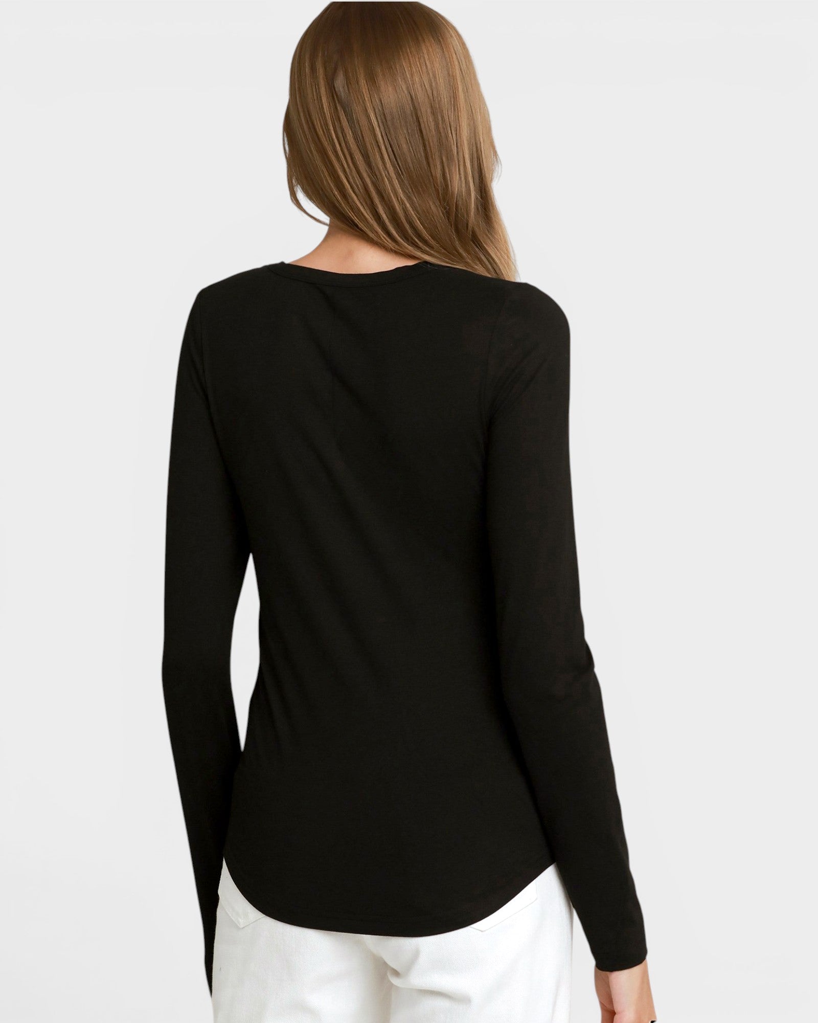 A woman viewed from the back wearing a black long-sleeve top and white pants against a light gray studio background.