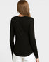 A woman viewed from the back wearing a black long-sleeve top and white pants against a light gray studio background.