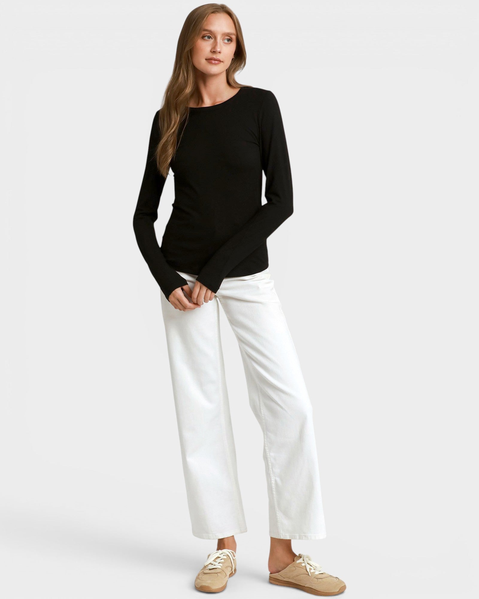 A woman modeling a black long-sleeve crewneck top with white wide-leg cropped pants and beige slip-on shoes, photographed against a light gray studio background.