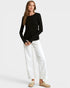 A woman modeling a black long-sleeve crewneck top with white wide-leg cropped pants and beige slip-on shoes, photographed against a light gray studio background.