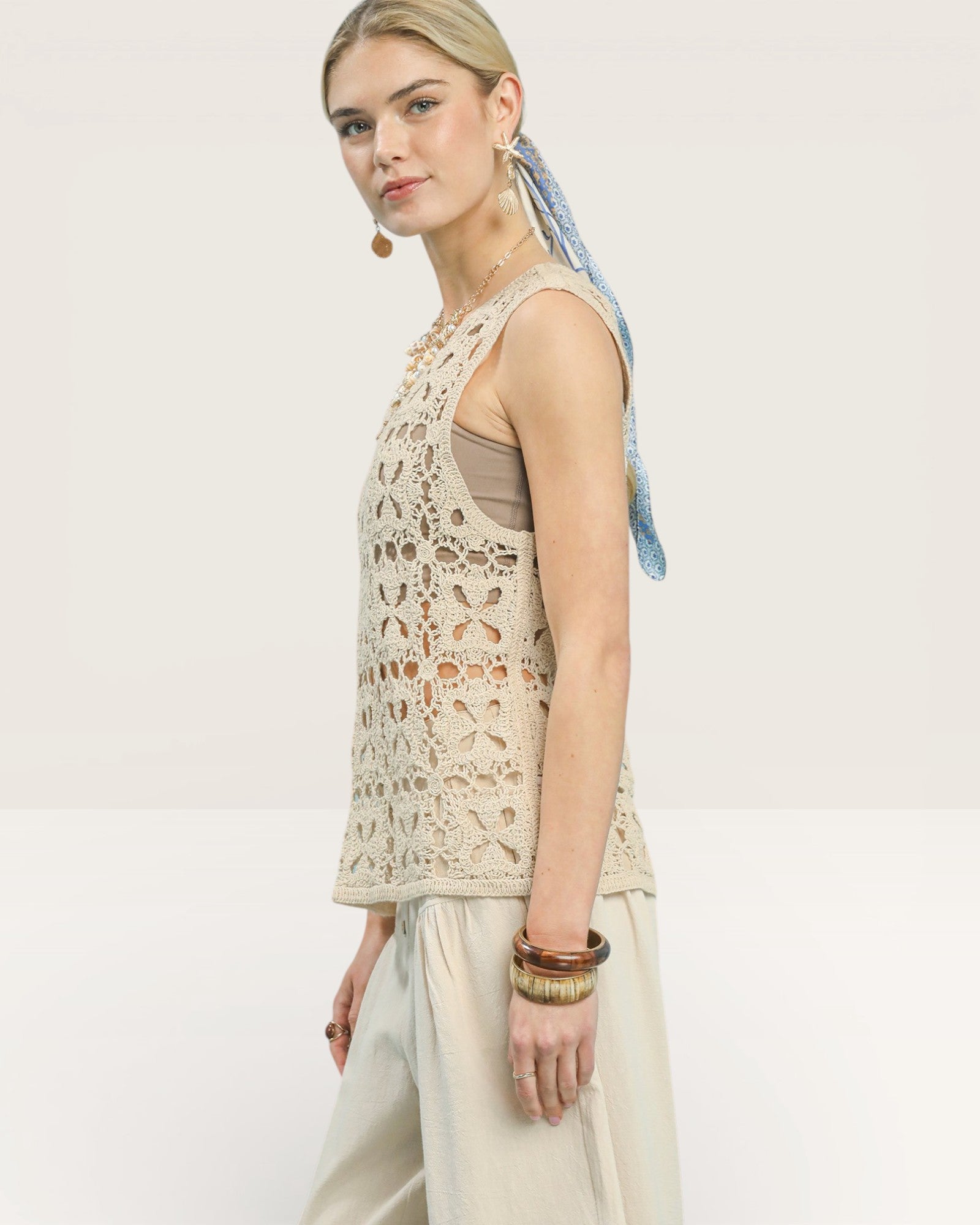 Woman wearing a beige sleeveless top with a pattern, white pants, and a headscarf on a plain background