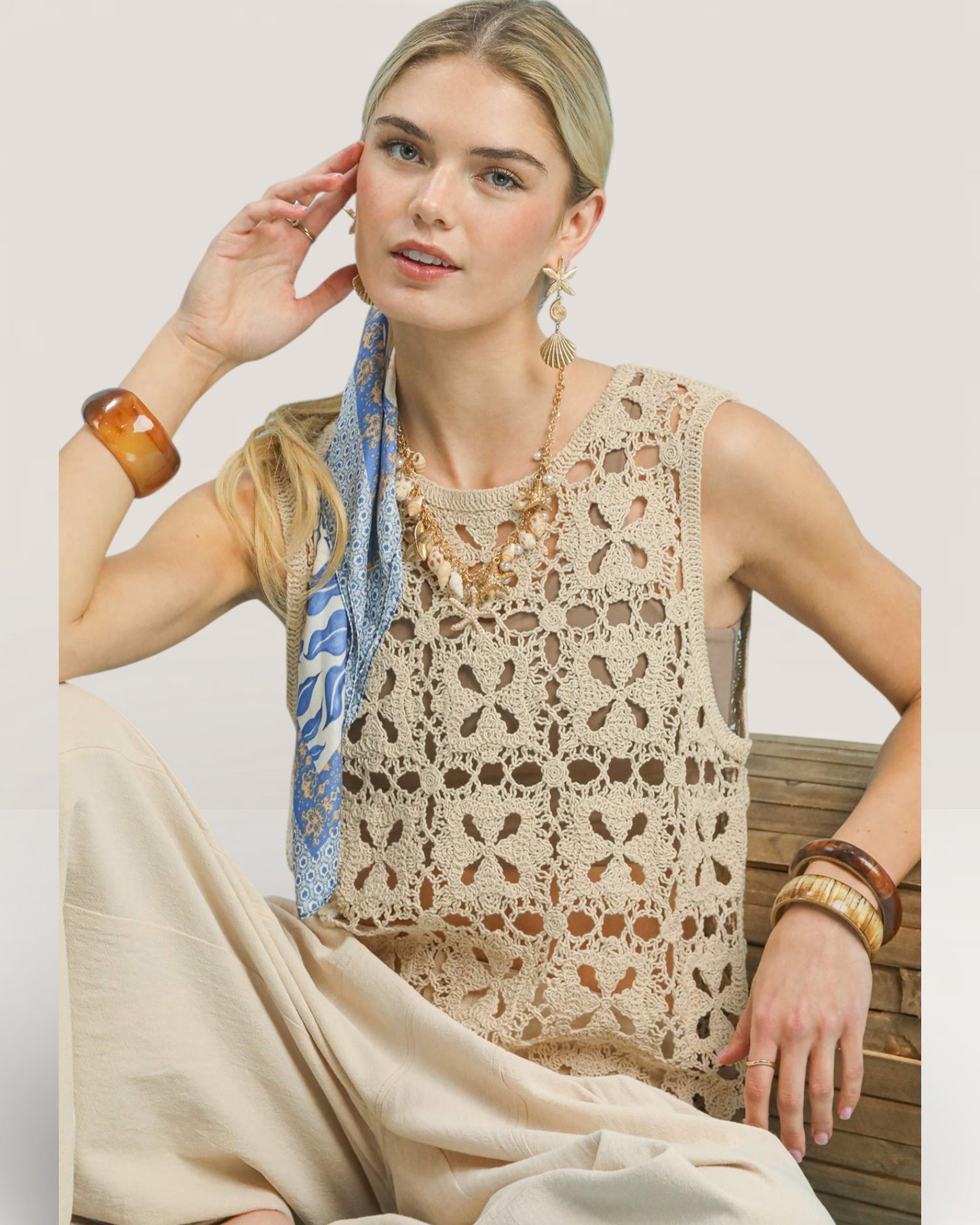 Woman wearing a beige crochet top and holding a blue scarf, sitting on a wooden bench.