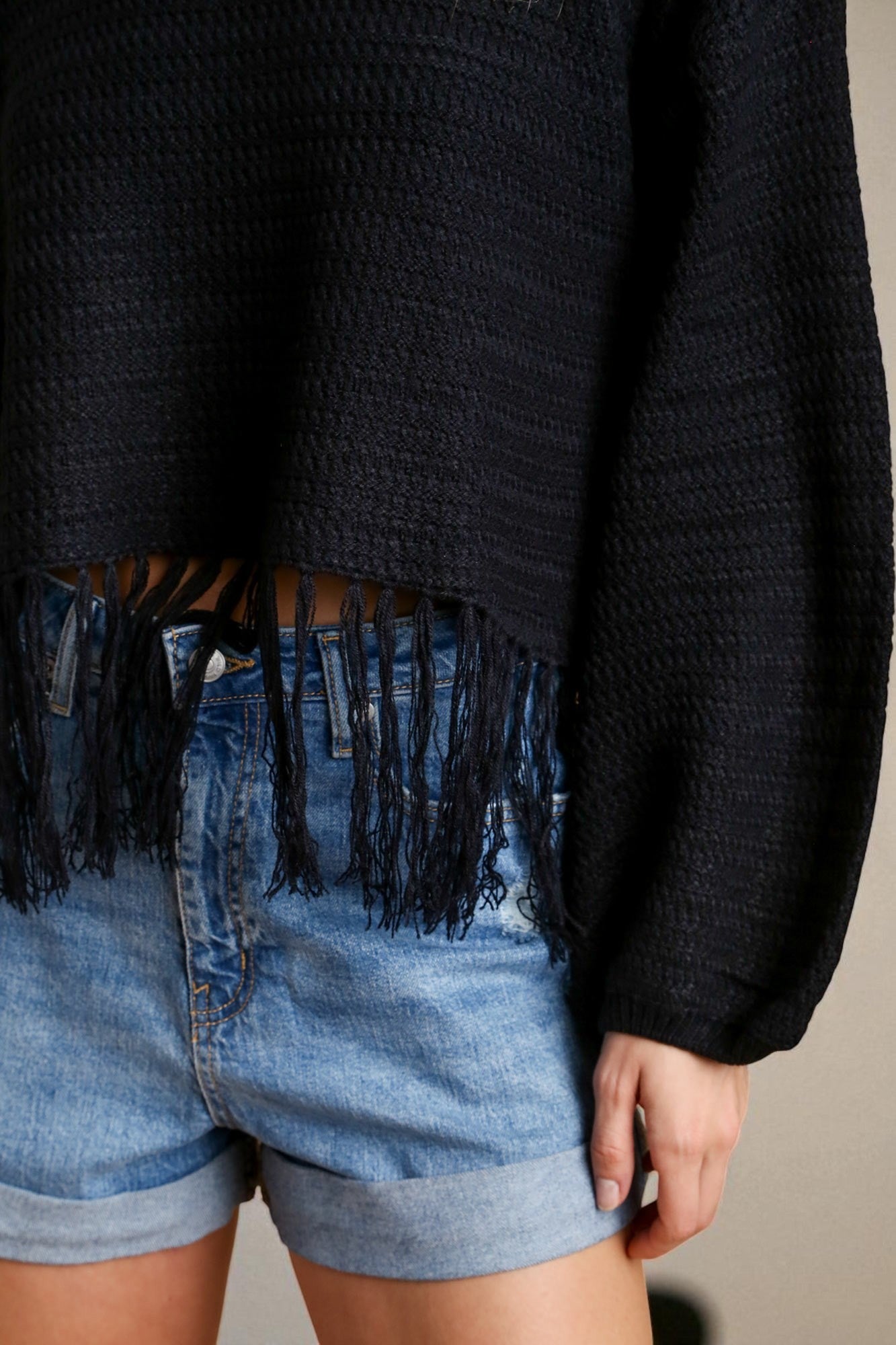 Person wearing a black sweater with fringe details and denim shorts on a neutral background