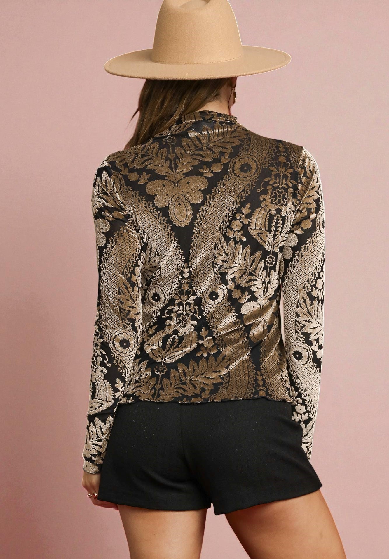 Person wearing a patterned jacket and black shorts with a beige hat on a pink background