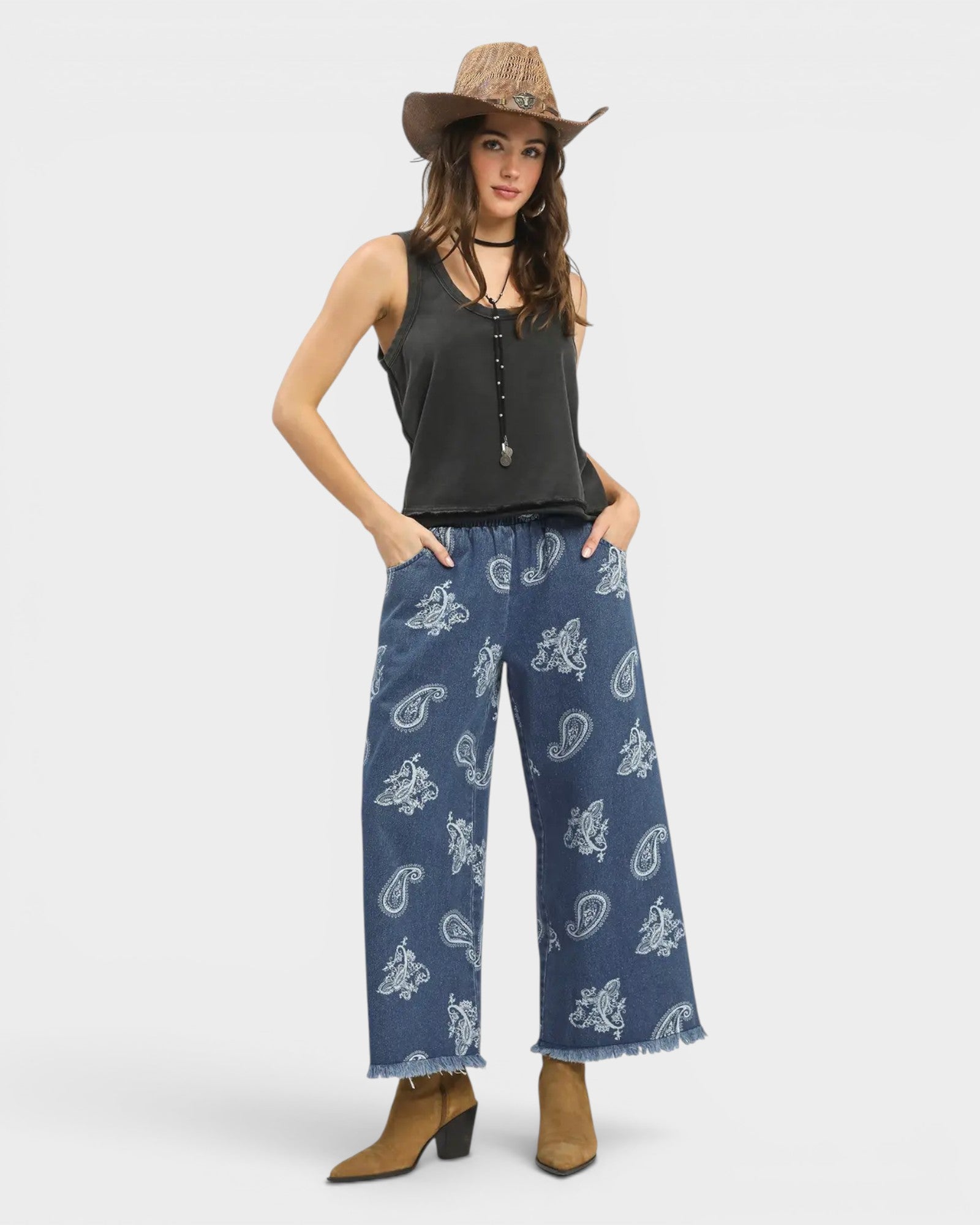 Woman wearing a black sleeveless top and blue paisley pants with a hat on a white background