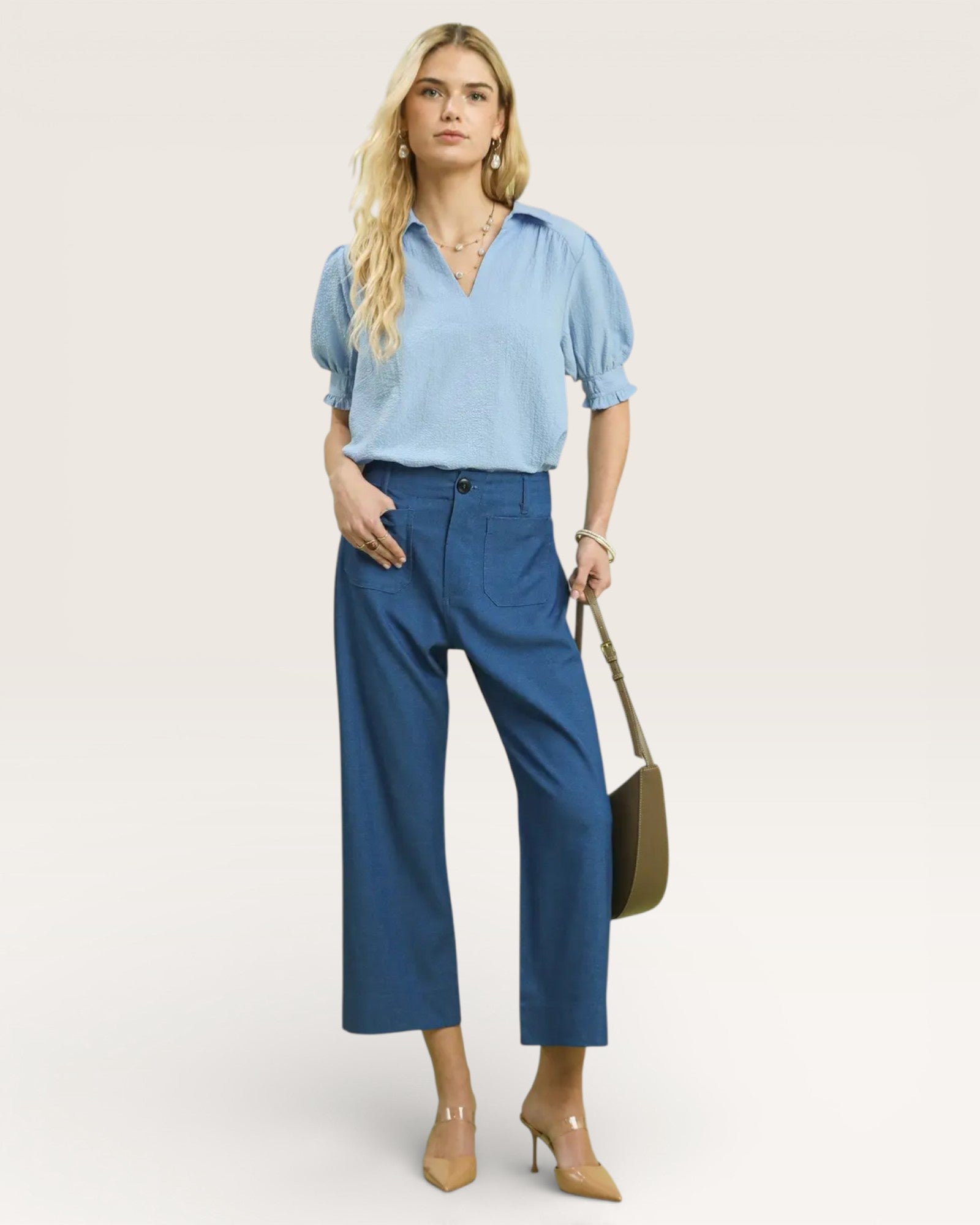 Woman wearing a light blue blouse and blue pants, holding a green handbag on a white background